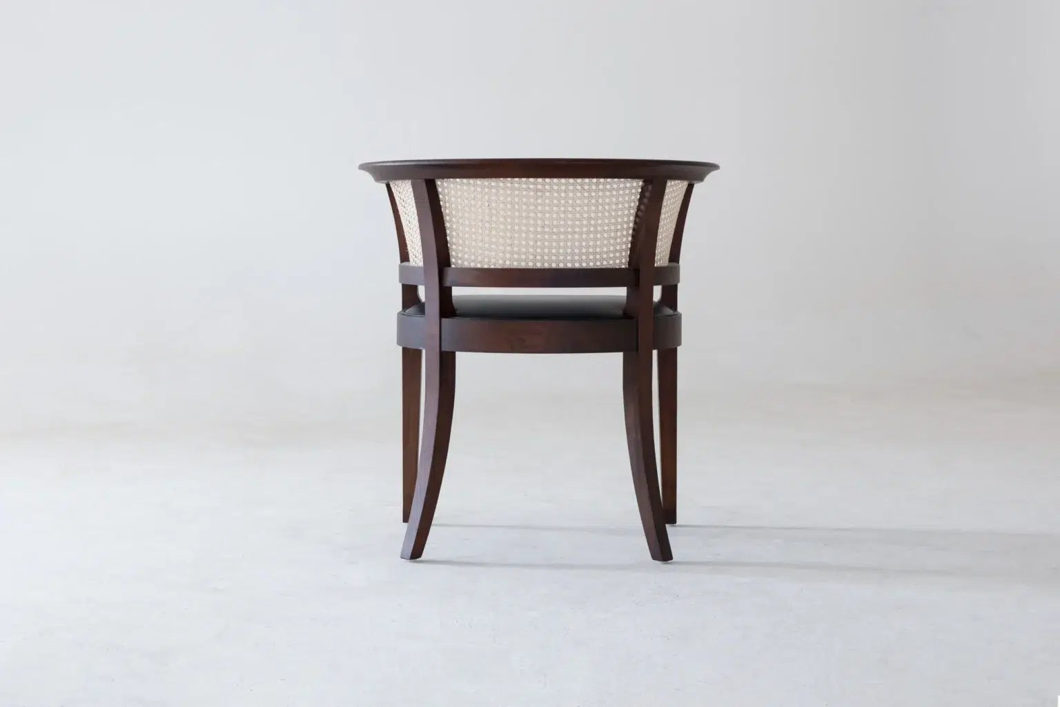 Back view A dark wooden chair with a curved backrest and woven cane panel, shown from behind, placed on a light-coloured floor against a plain, light grey background.