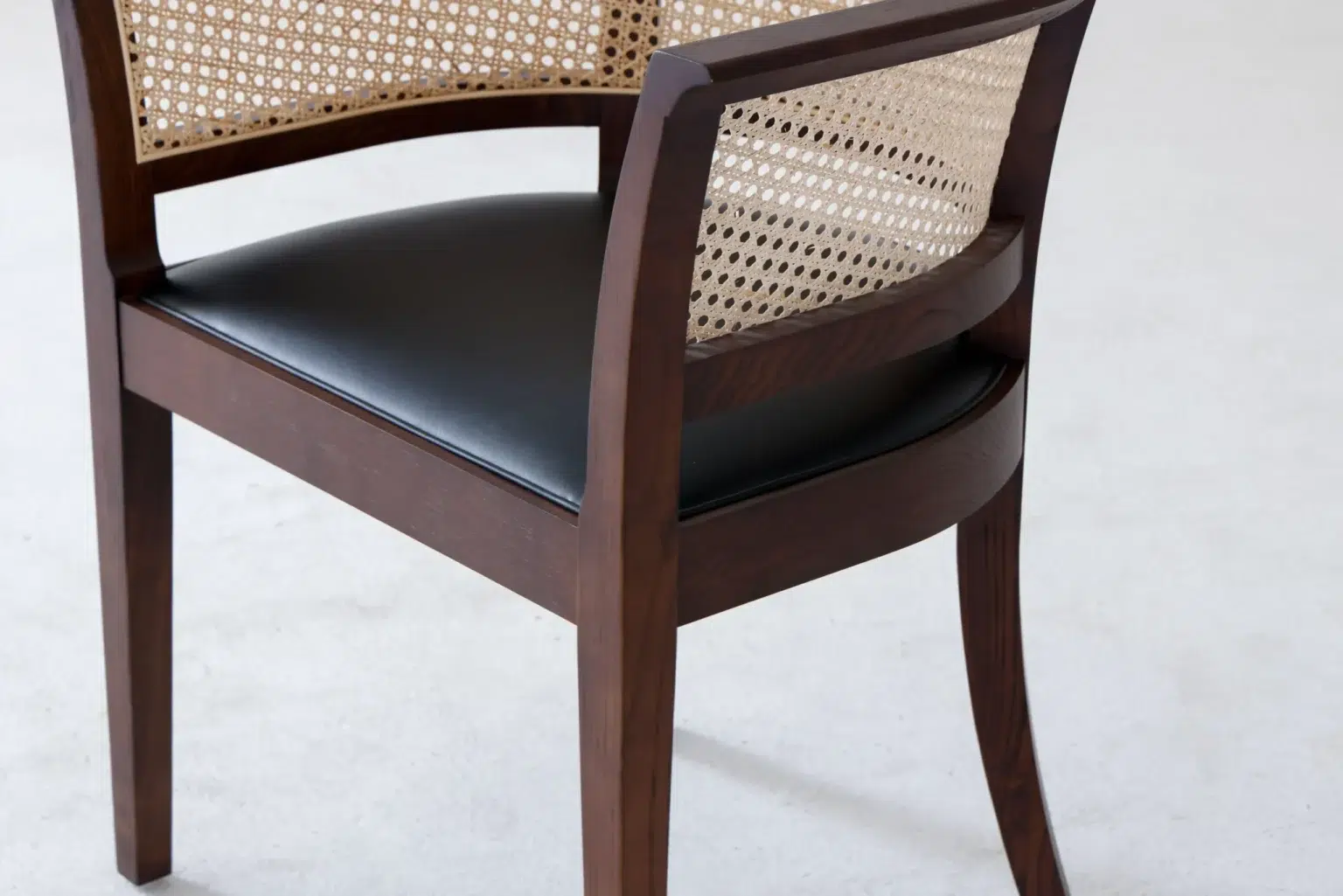Detail display1 A wooden chair with a curved cane backrest and a black cushioned seat, shown on a light-coloured floor.