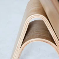Close-up of a modern, bent plywood furniture piece with smooth curves, light wood grain, and a layered construction, set against a plain, light background.