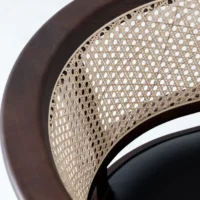 Close-up of a chair with a dark wood frame and a curved backrest featuring a woven cane pattern. The seat appears to be solid and black, contrasting with the light-coloured cane.