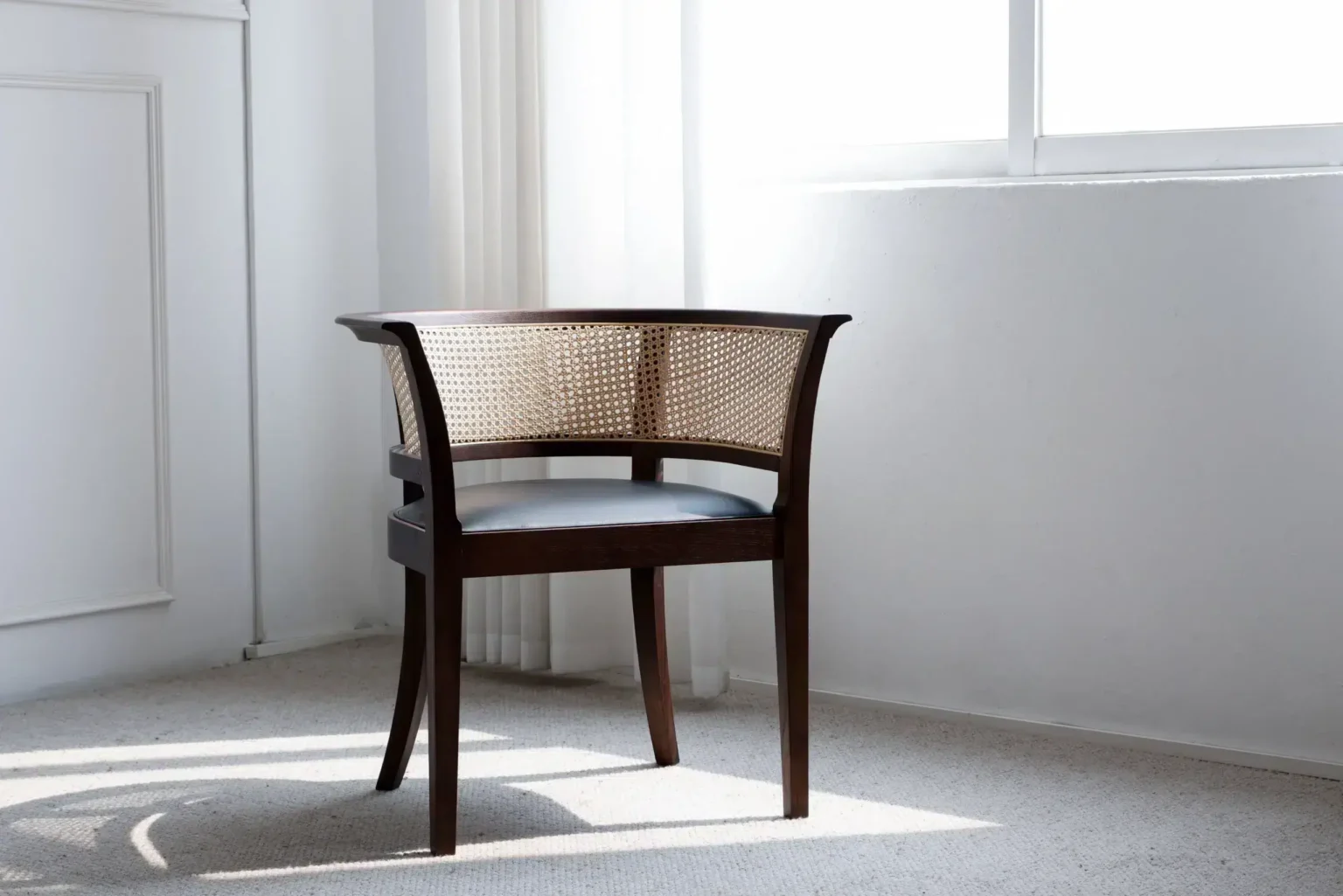 Display image A wooden chair with a woven rattan backrest and a black seat cushion sits in a bright, minimalist room with carpeted flooring and sunlight streaming through a nearby window.