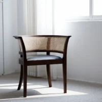 A wooden chair with a woven rattan backrest and a black seat cushion sits in a bright, minimalist room with carpeted flooring and sunlight streaming through a nearby window.