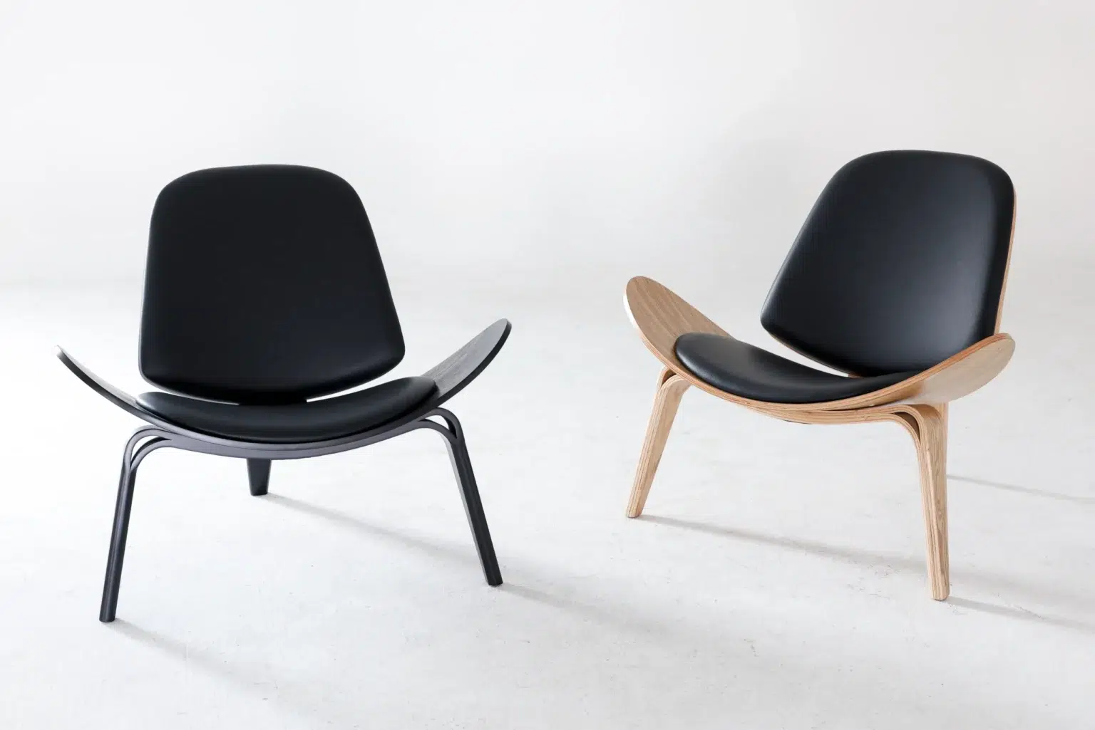 Display image Two modern lounge chairs with curved seats and backs. One chair has black legs and frame, whilst the other has light wooden legs and frame. Both chairs have black cushions and are set against a plain white background.