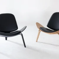 Two modern lounge chairs with curved seats and backs. One chair has black legs and frame, whilst the other has light wooden legs and frame. Both chairs have black cushions and are set against a plain white background.