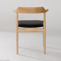 Minimalist wooden chair with light wood frame, curved backrest, and black cushioned seat. The chair is photographed against a plain white background and is shown from the back.
