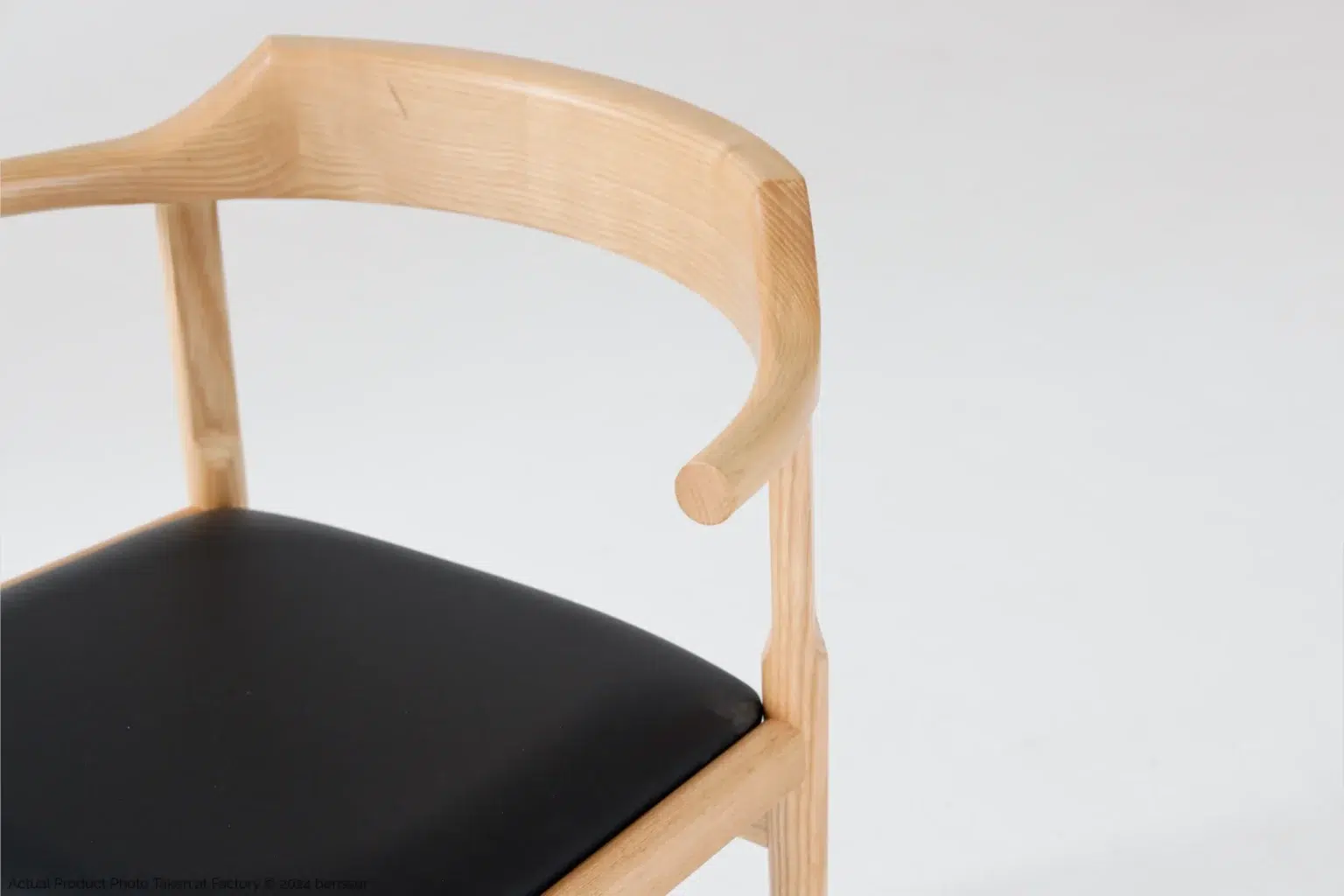 Hans Wegner_Final Chair_Replica_Microfiber_Natural_Ash_Detail_1 A close-up of a modern wooden chair with a curved backrest and a black cushioned seat, set against a plain white background.