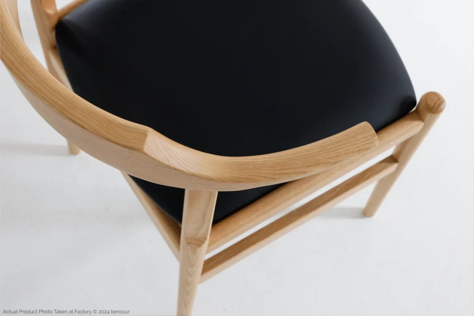 Hans Wegner_Final Chair_Replica_Microfiber_Natural_Ash_Detail_2 A close-up view of a wooden chair with a smooth, curved backrest and a black cushioned seat, placed on a white surface.