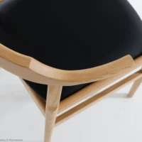 A close-up view of a wooden chair with a smooth, curved backrest and a black cushioned seat, placed on a white surface.