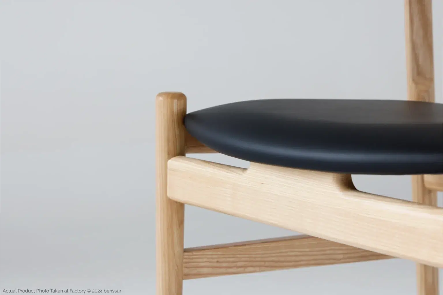 Hans Wegner_Final Chair_Replica_Microfiber_Natural_Ash_Detail_3 Close-up of a modern wooden chair with light-coloured legs and frame, featuring a smooth, curved black seat cushion against a plain light grey background.