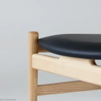 Close-up of a modern wooden chair with light-coloured legs and frame, featuring a smooth, curved black seat cushion against a plain light grey background.