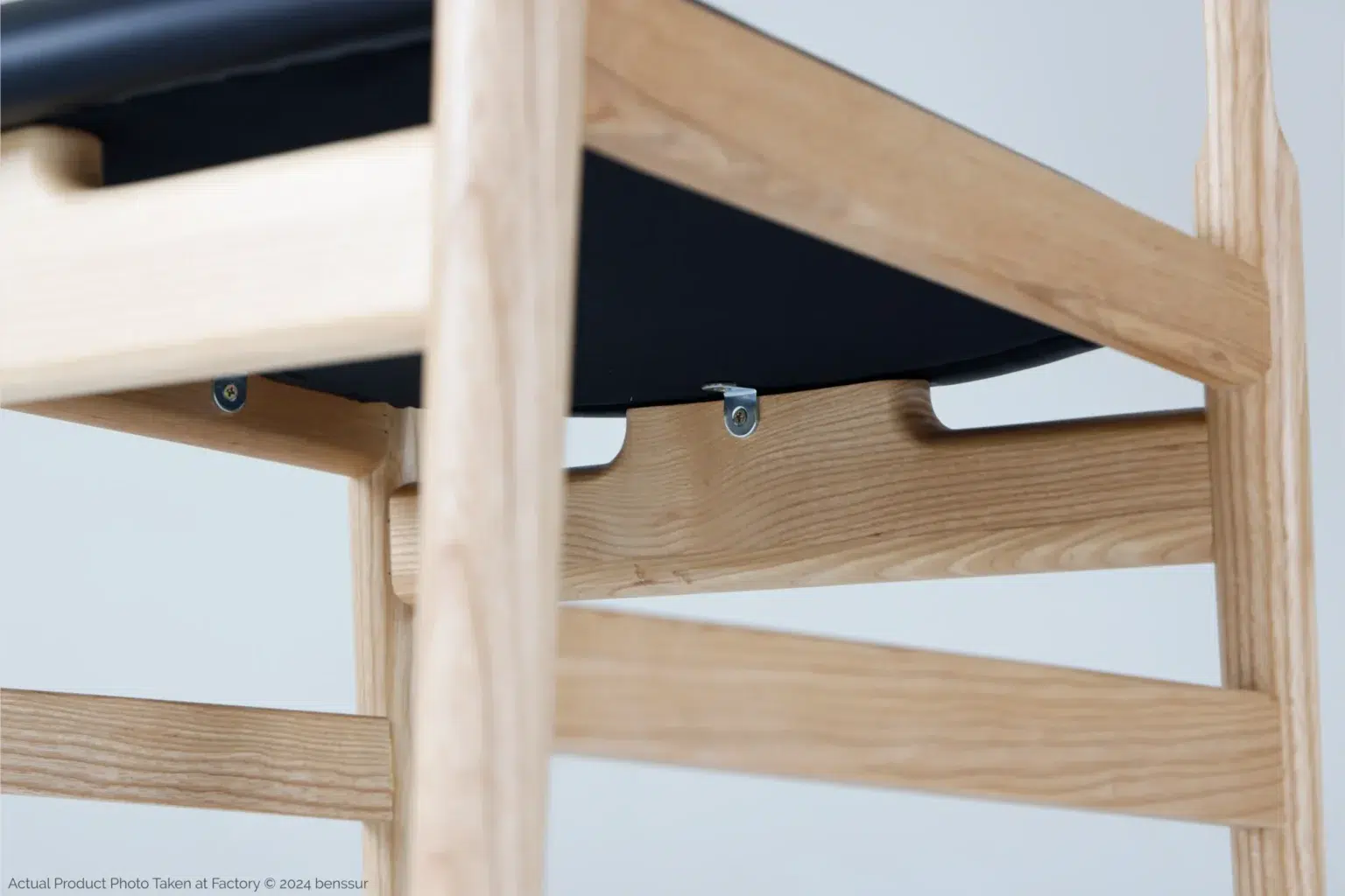 Hans Wegner_Final Chair_Replica_Microfiber_Natural_Ash_Detail_4 Close-up view of a wooden chair frame with a black seat. The light wood has a smooth finish, and metal fittings are visible beneath the seat. The background is plain and light-coloured.