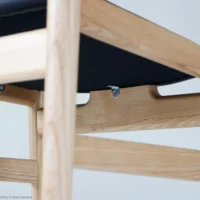 Close-up view of a wooden chair frame with a black seat. The light wood has a smooth finish, and metal fittings are visible beneath the seat. The background is plain and light-coloured.