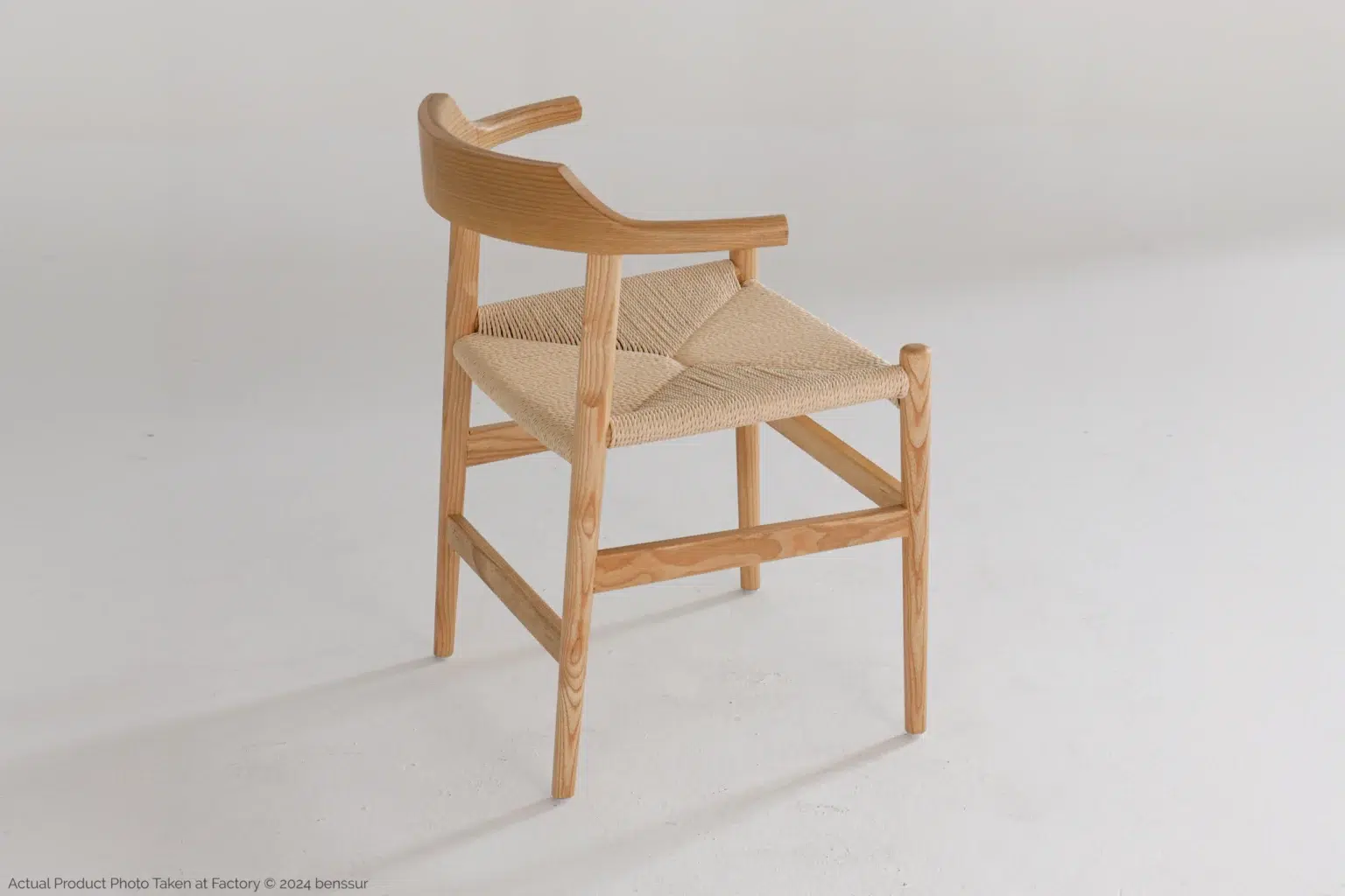 Hans Wegner_Final Chair_Replica_Paper Rope_Natural_Ash_Back Right A wooden chair with a woven seat and a curved, low backrest is positioned on a light-coloured floor against a plain white background.