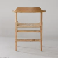 A minimalist wooden chair with a curved backrest and woven seat, set against a plain light background. The chair has clean lines and a natural finish.