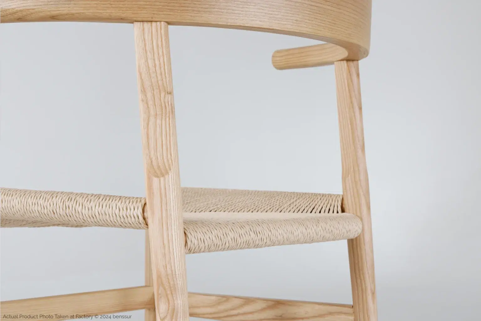 Hans Wegner_Final Chair_Replica_Paper Rope_Natural_Ash_Detail_1 Close-up of a light wooden chair with a woven seat and smooth, rounded edges, shown against a plain, light background. The craftsmanship and texture of the materials are clearly visible.