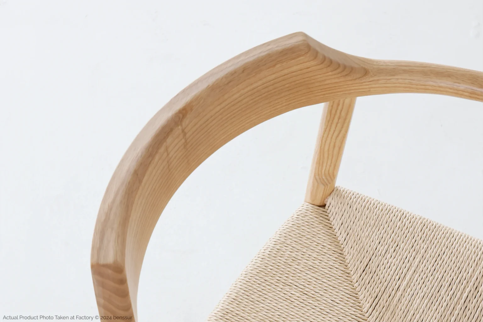 Hans Wegner_Final Chair_Replica_Paper Rope_Natural_Ash_Detail_2 A close-up, angled view of a wooden chair with a curved, smooth backrest and a woven, natural fibre seat against a plain white background.
