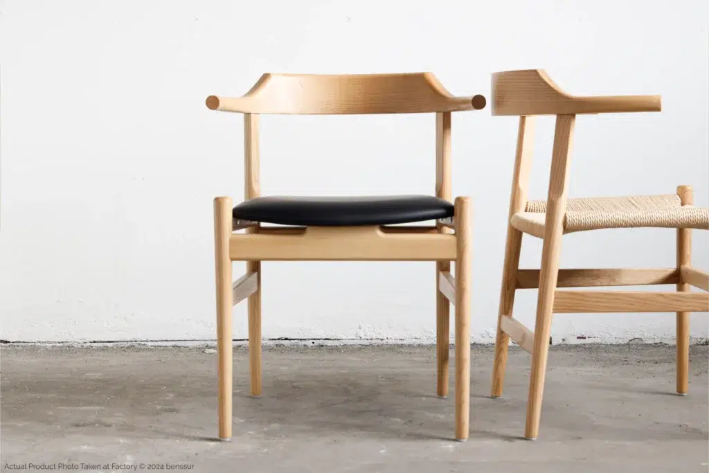 Two modern wooden chairs with curved backrests; one has a black cushioned seat, and the other has a woven seat. Both chairs are on a concrete floor against a plain white wall.