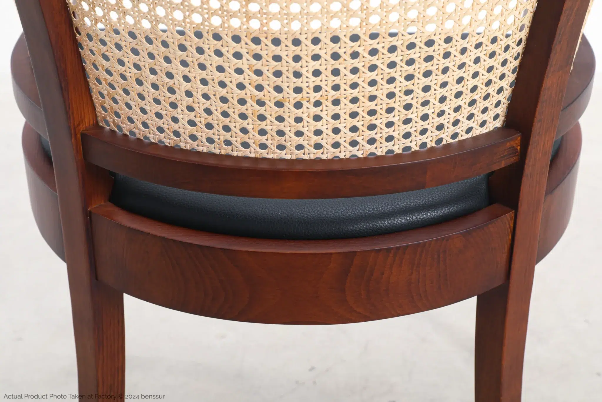 Close-up view of the back of a wooden chair with a woven cane backrest and a black cushioned seat, set against a light background.