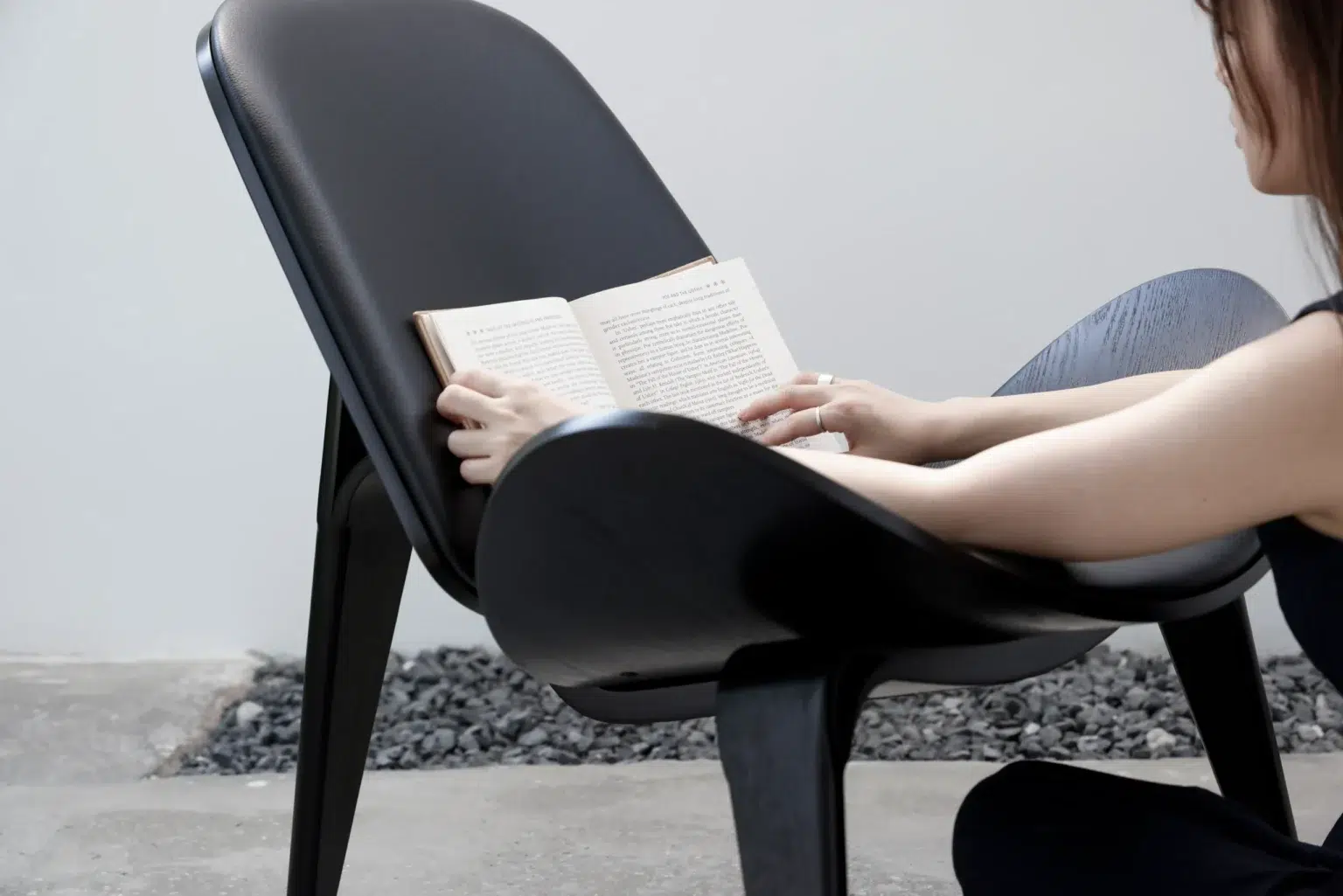 Outdoor30 A person sitting in a modern black chair, reading an open book, with a minimalist background featuring grey pebbles and a concrete floor.