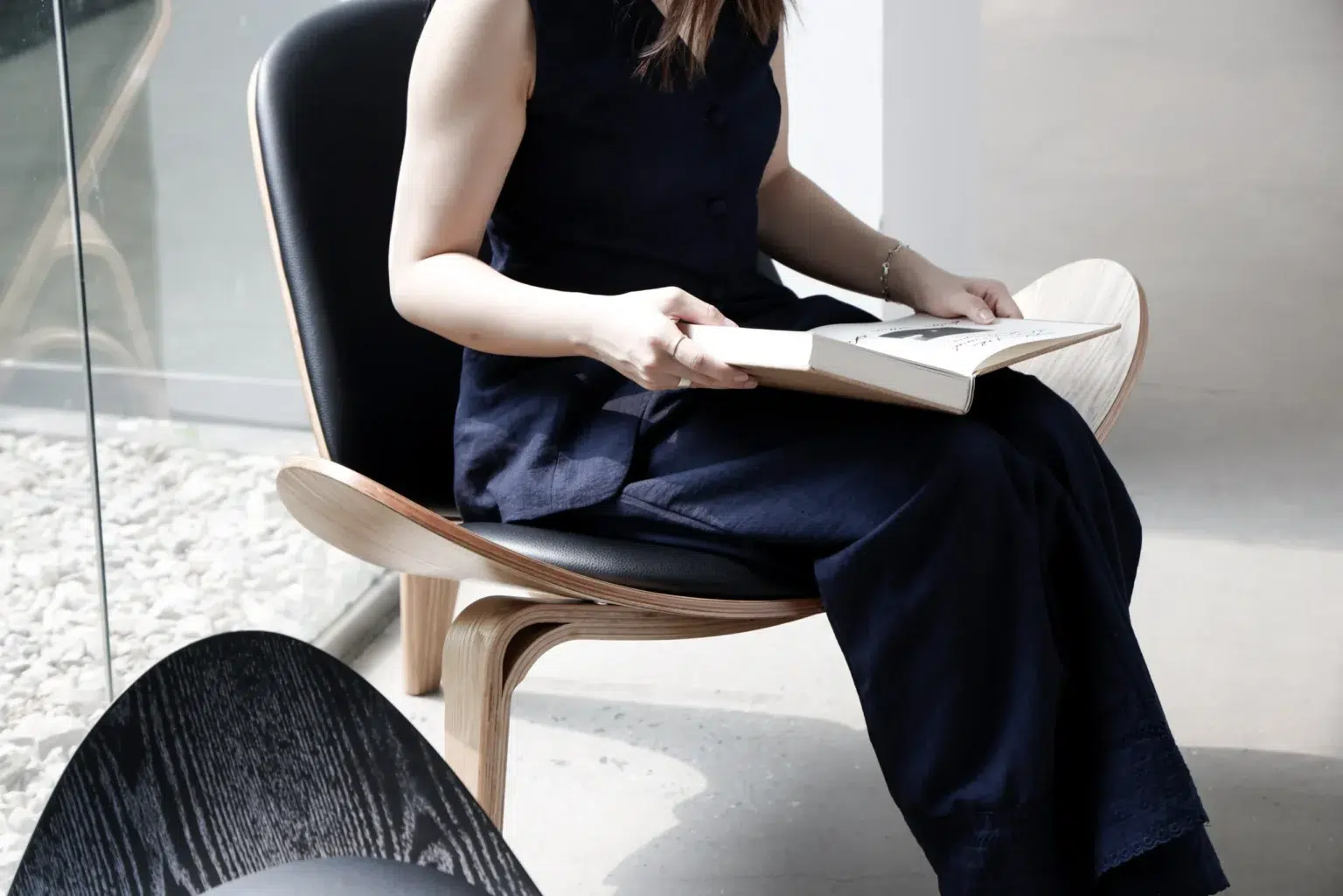 Outdoor31 A woman in a sleeveless navy outfit sits on a modern wooden chair near a window, holding an open book in her lap. Bright natural light fills the minimalist, contemporary room.