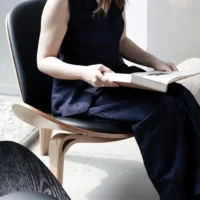 A woman in a sleeveless navy outfit sits on a modern wooden chair near a window, holding an open book in her lap. Bright natural light fills the minimalist, contemporary room.