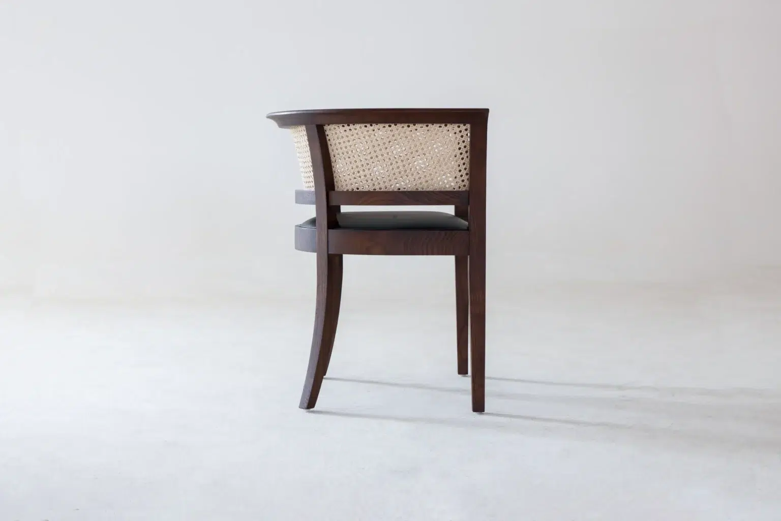 Side view A wooden chair with a woven cane backrest and dark legs, placed on a plain white floor against a light grey wall, facing away from the camera.