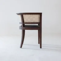 A wooden chair with a woven cane backrest and dark legs, placed on a plain white floor against a light grey wall, facing away from the camera.