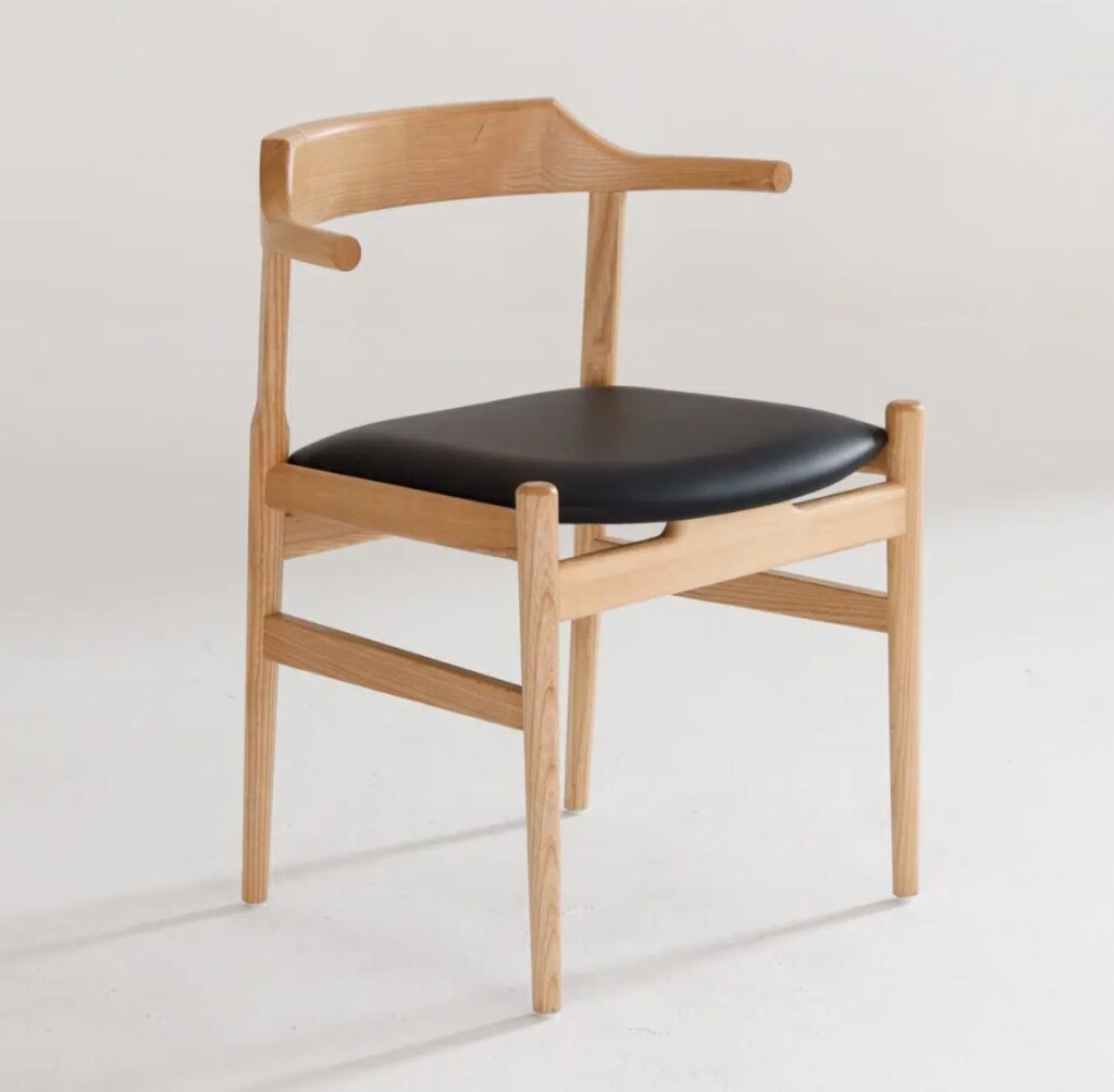 A modern wooden chair with angled legs, a curved backrest, and a black cushioned seat, placed on a plain light-coloured surface.