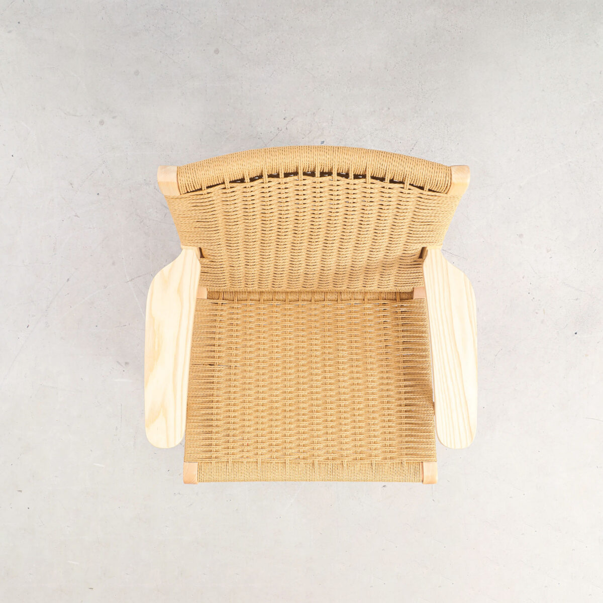 Easy Chair_B11 Top view of a woven rattan chair with wooden armrests on a light grey concrete floor, showing the intricate weave pattern and natural materials.