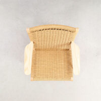 Top view of a woven rattan chair with wooden armrests on a light grey concrete floor, showing the intricate weave pattern and natural materials.
