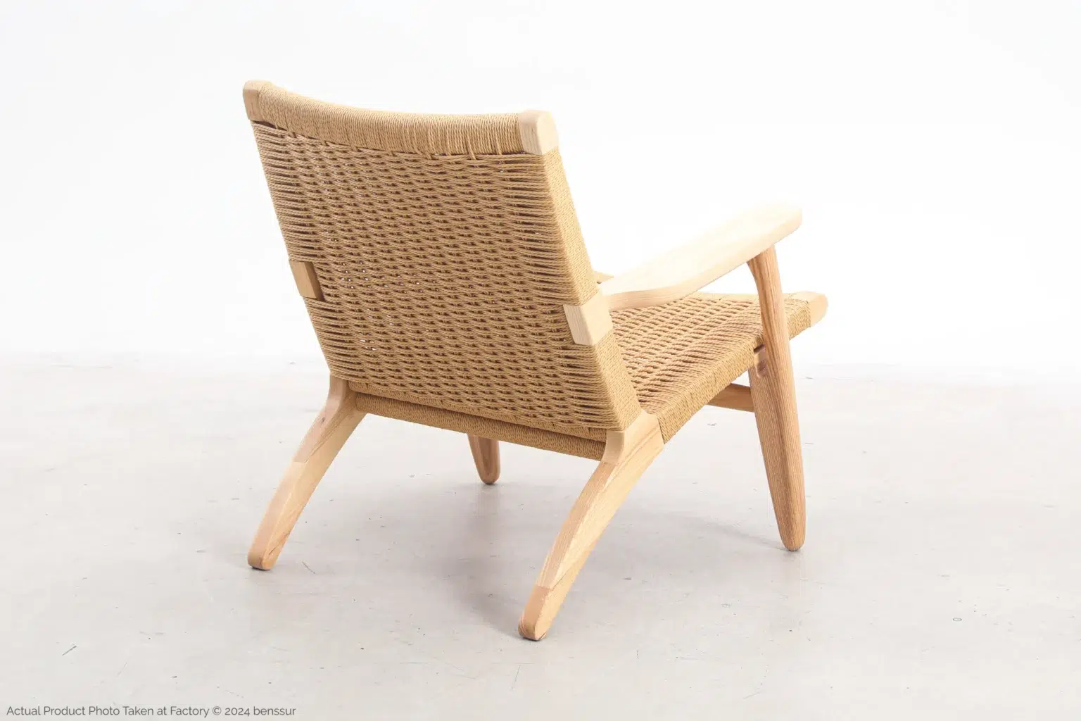 Hans Wegner_CH25 Easy Chair_Replica_Paper Rope_Natural_Ash_Back Right A wooden lounger with a woven backrest and seat, featuring light-coloured wood and angled legs, is shown on a light floor against a plain white background. The chair is viewed from behind and slightly to the side.