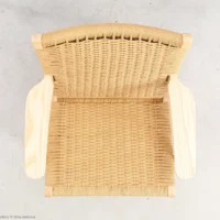Top view of a woven rattan chair with light wooden armrests on a light-coloured floor. The chair has a natural, minimalist design and a rectangular seat and backrest.