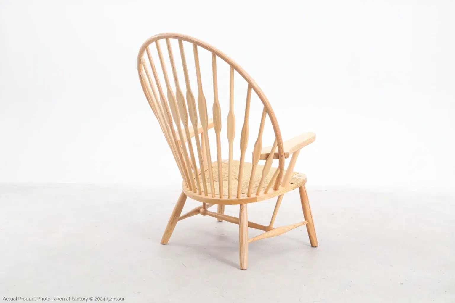 Hans Wegner_Peacock Chair_Replica_Paper Rope_Natural_Ash_Back Right A wooden Windsor-style chair with a high, curved spindle back and flat armrests, set on a light-coloured floor against a plain white background.