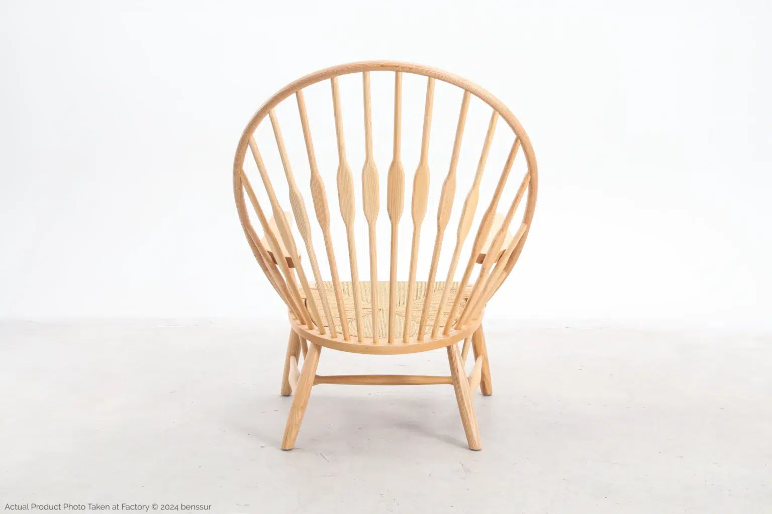 Hans Wegner_Peacock Chair_Replica_Paper Rope_Natural_Ash_Back A light wood chair with a round, high spindle back and angled legs sits on a plain, light-coloured floor against a white background. The seat appears woven and the overall design is minimalist and modern.
