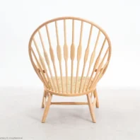 A light wood chair with a round, high spindle back and angled legs sits on a plain, light-coloured floor against a white background. The seat appears woven and the overall design is minimalist and modern.
