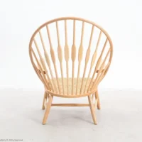 A light wood chair with a round, high spindle back and angled legs sits on a plain, light-coloured floor against a white background. The seat appears woven and the overall design is minimalist and modern.