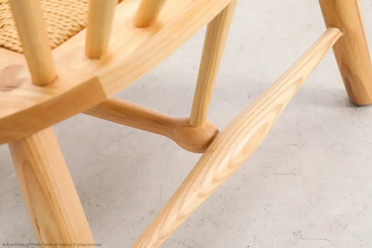 Hans Wegner_Peacock Chair_Replica_Paper Rope_Natural_Ash_Detail_1 Close-up of a light wooden chair leg and stretcher with visible wood grain, showcasing craftsmanship and joinery. The seat appears woven, and the chair is on a light-coloured floor.