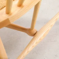 Close-up of a light wooden chair leg and stretcher with visible wood grain, showcasing craftsmanship and joinery. The seat appears woven, and the chair is on a light-coloured floor.