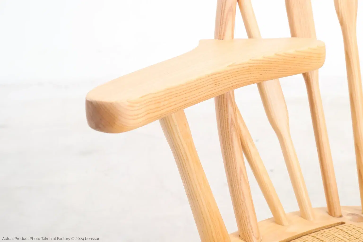 Hans Wegner_Peacock Chair_Replica_Paper Rope_Natural_Ash_Detail_3 Close-up of a light wooden chair with smooth curves and a natural finish, showing the armrest and part of the backrest against a plain, white background.