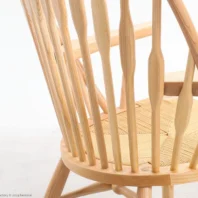 Close-up of a light wooden chair with vertical spindle backrest, curved armrests, and a woven seat, set against a plain white background.