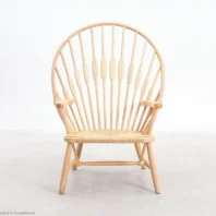 A light wood Windsor-style chair with a rounded, high spindle back and woven seat, sitting on a plain, light-coloured floor against a white background.