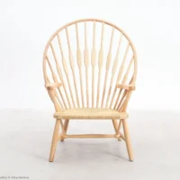 A light wood Windsor-style chair with a rounded, high spindle back and woven seat, sitting on a plain, light-coloured floor against a white background.