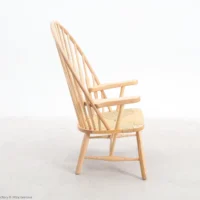 Side view of a wooden armchair with a high, curved spindle back, woven seat, and angled legs, set against a plain white background.