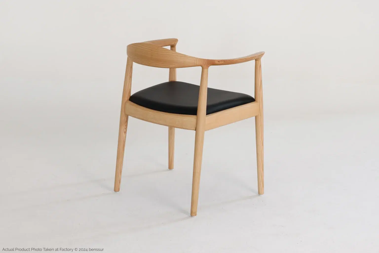 Hans Wegner_Round Chair_Replica_Microfiber_Natural_Ash_Back Right A modern wooden chair with a curved backrest and black cushioned seat is positioned on a plain white background, angled slightly to show its side and back.