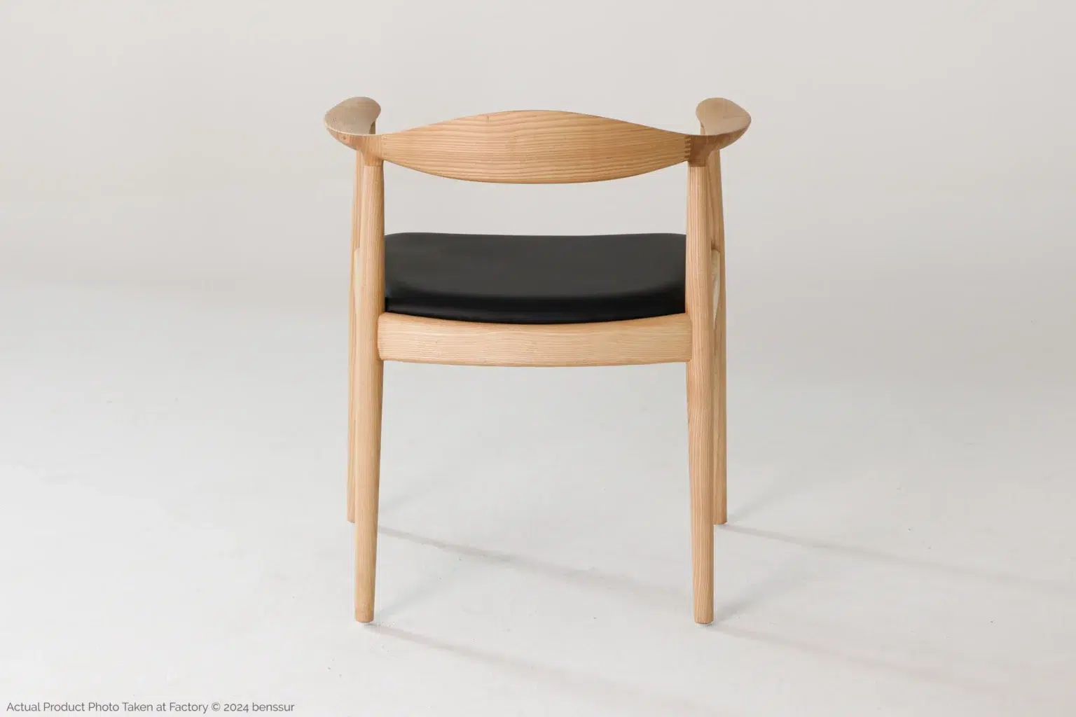 Hans Wegner_Round Chair_Replica_Microfiber_Natural_Ash_Back A wooden chair with a curved backrest, armrests, and a black cushioned seat is shown against a plain white background. The design is simple and modern, with smooth, light-coloured wood.