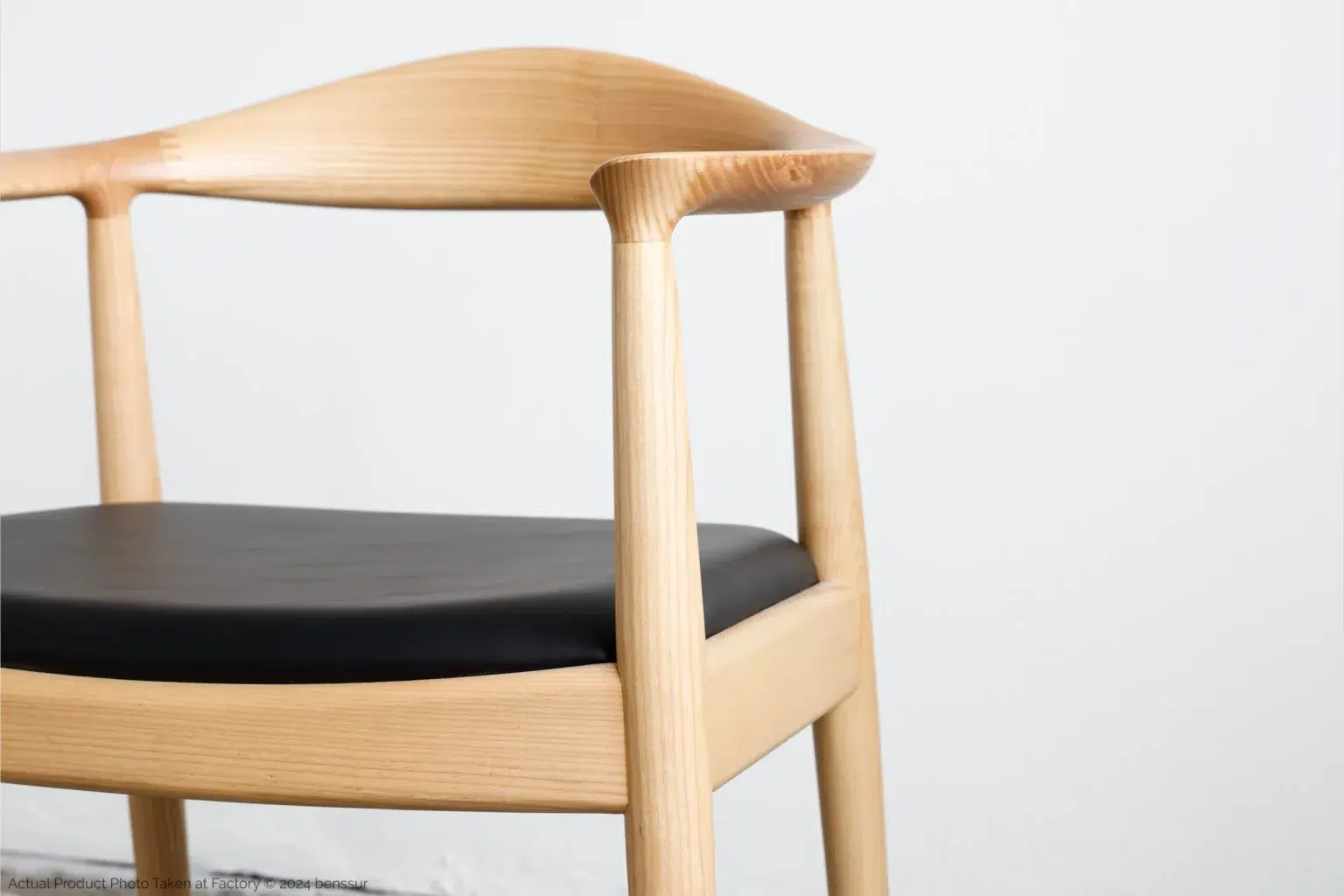 Hans Wegner_Round Chair_Replica_Microfiber_Natural_Ash_Detail_2 A close-up of a modern wooden chair with a curved backrest and black cushioned seat, set against a plain white background.