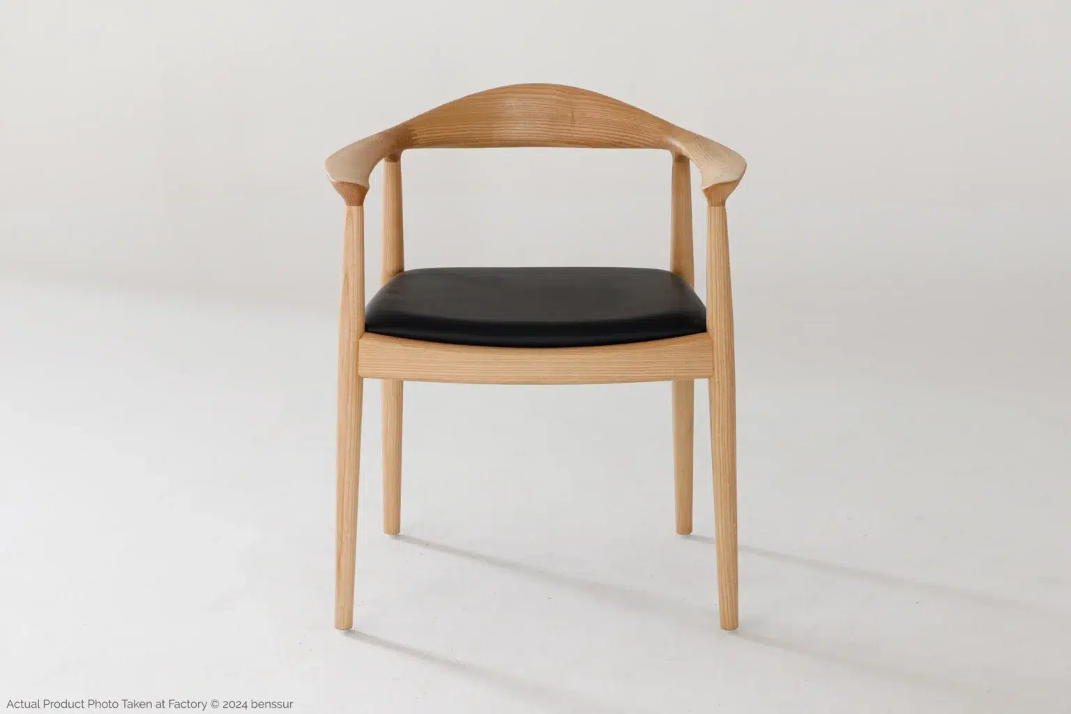 Hans Wegner_Round Chair_Replica_Microfiber_Natural_Ash_Front A wooden chair with curved armrests and a black cushioned seat, set against a plain white background. The design is minimalist and modern.