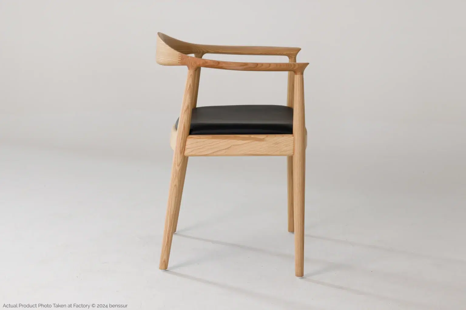 Hans Wegner_Round Chair_Replica_Microfiber_Natural_Ash_Right A modern wooden chair with a curved backrest and black cushioned seat, shown in profile against a plain white background. The design is minimalist with clean lines and light wood tones.