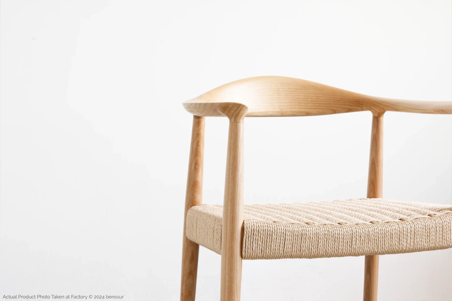 Hans Wegner_Round Chair_Replica_Paper Rope_Natural_Ash_Detail_1 Minimalist wooden chair with a curved backrest and woven seat, set against a plain white background. The design highlights smooth, light-coloured wood and simple, elegant lines.
