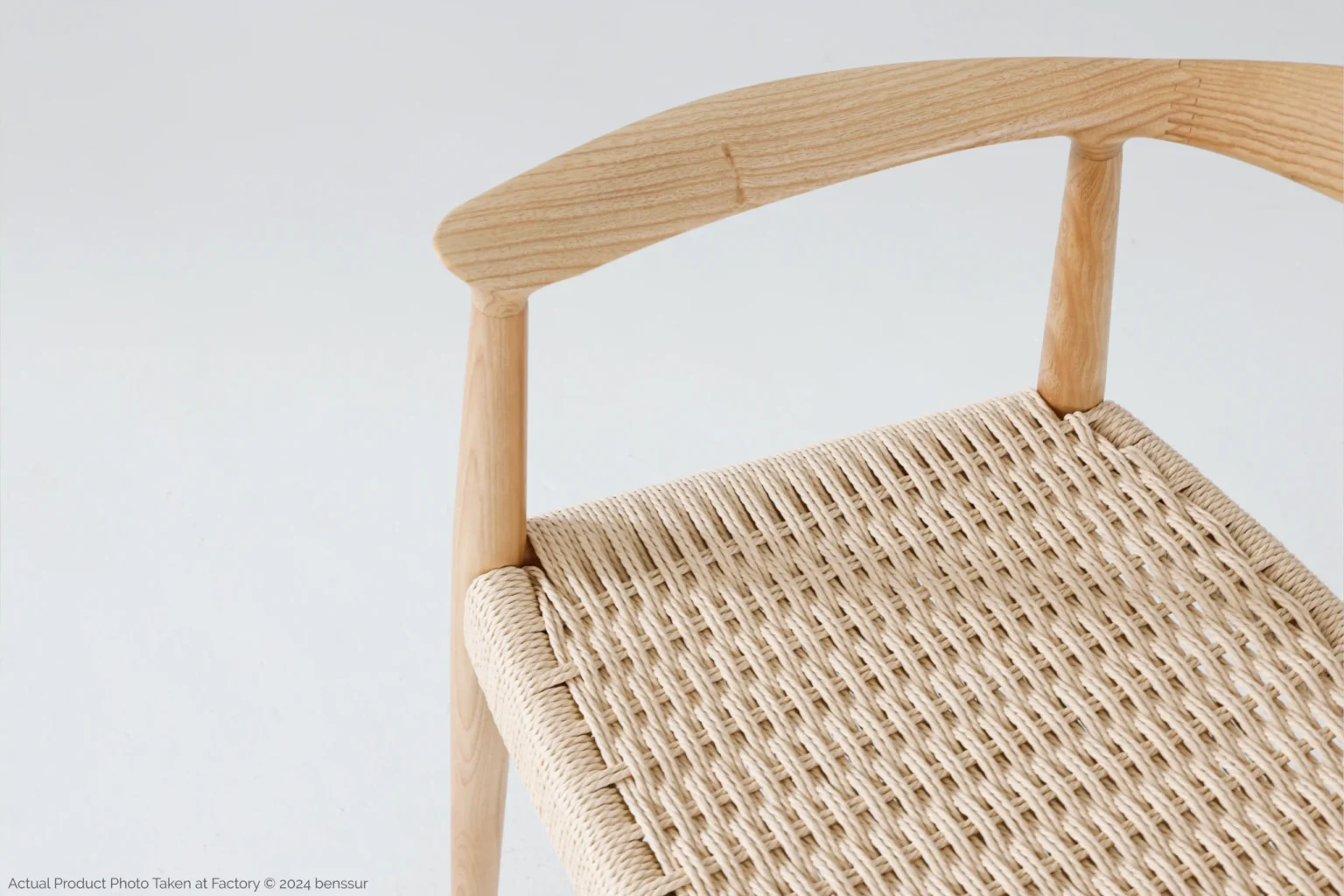 Hans Wegner_Round Chair_Replica_Paper Rope_Natural_Ash_Detail_2 A close-up of a wooden chair with a woven seat and a curved wooden backrest, shown against a plain light background. The craftsmanship of the natural wood and woven pattern is clearly visible.